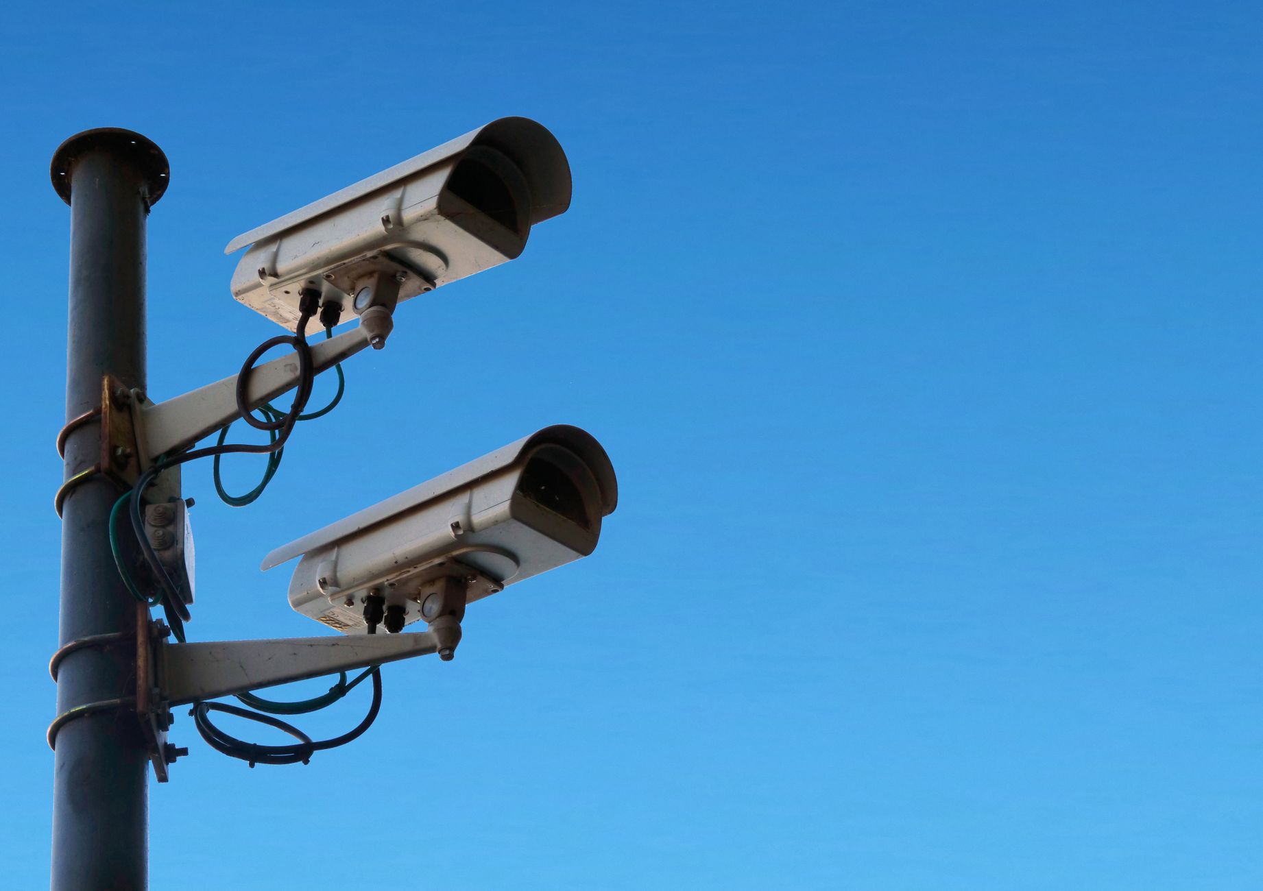Deux caméras de surveillance sous boîtier fixées à un mât métallique, avec des câbles visibles, devant un ciel bleu dégagé.