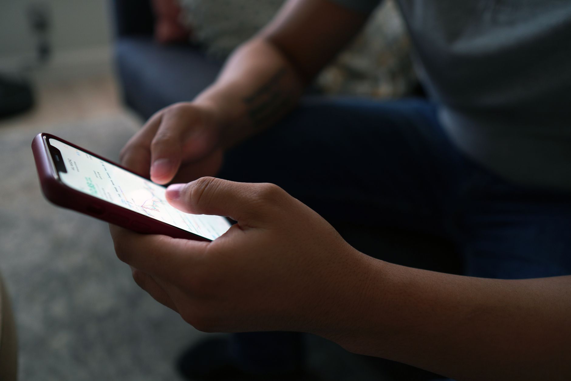 Mains d’une personne tenant un smartphone à étui rouge foncé, écran allumé, dans un intérieur aux tons sombres.