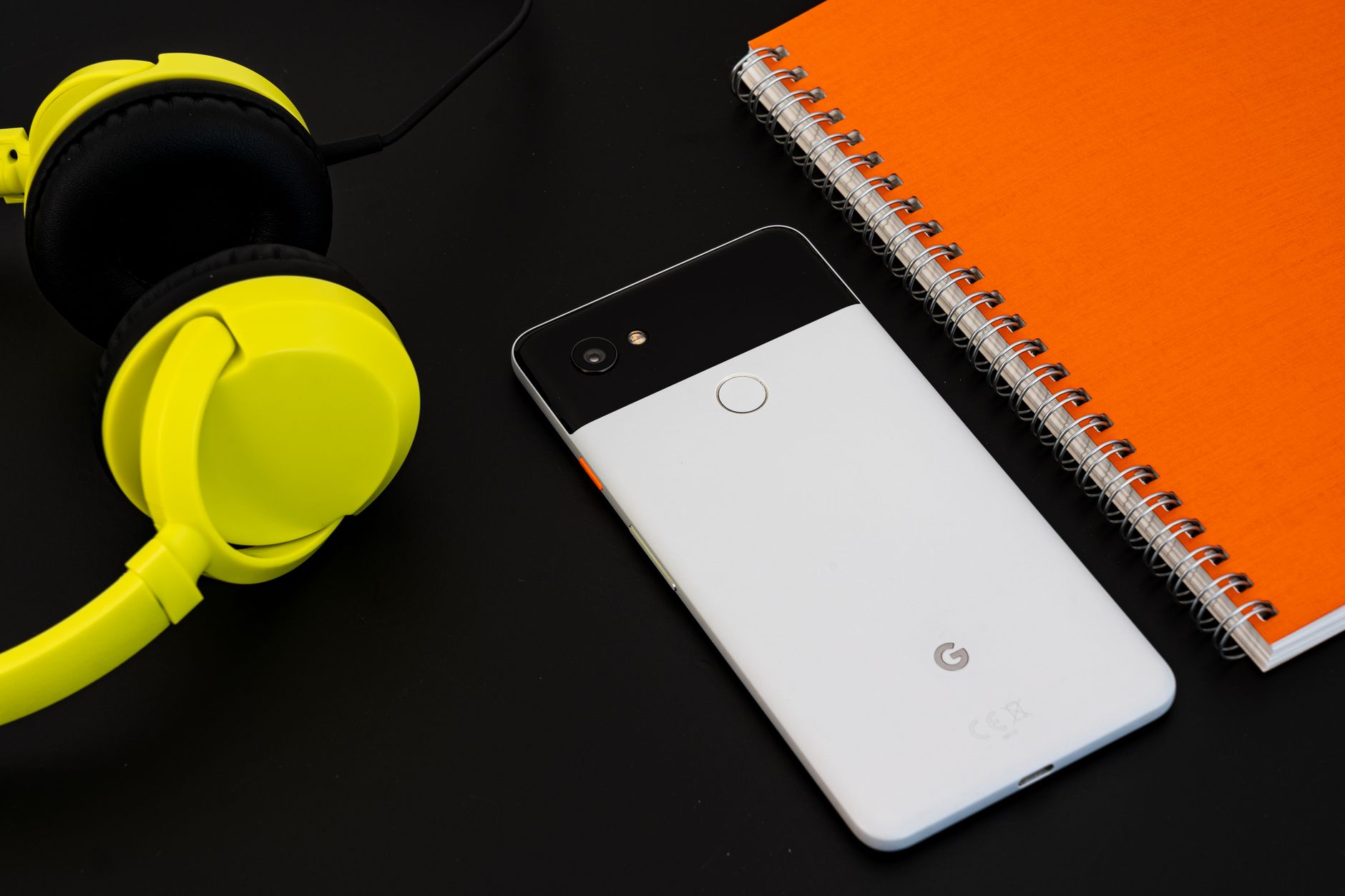 Smartphone noir et blanc posé face arrière entre un casque audio jaune fluo et un cahier à spirale orange sur un fond sombre.