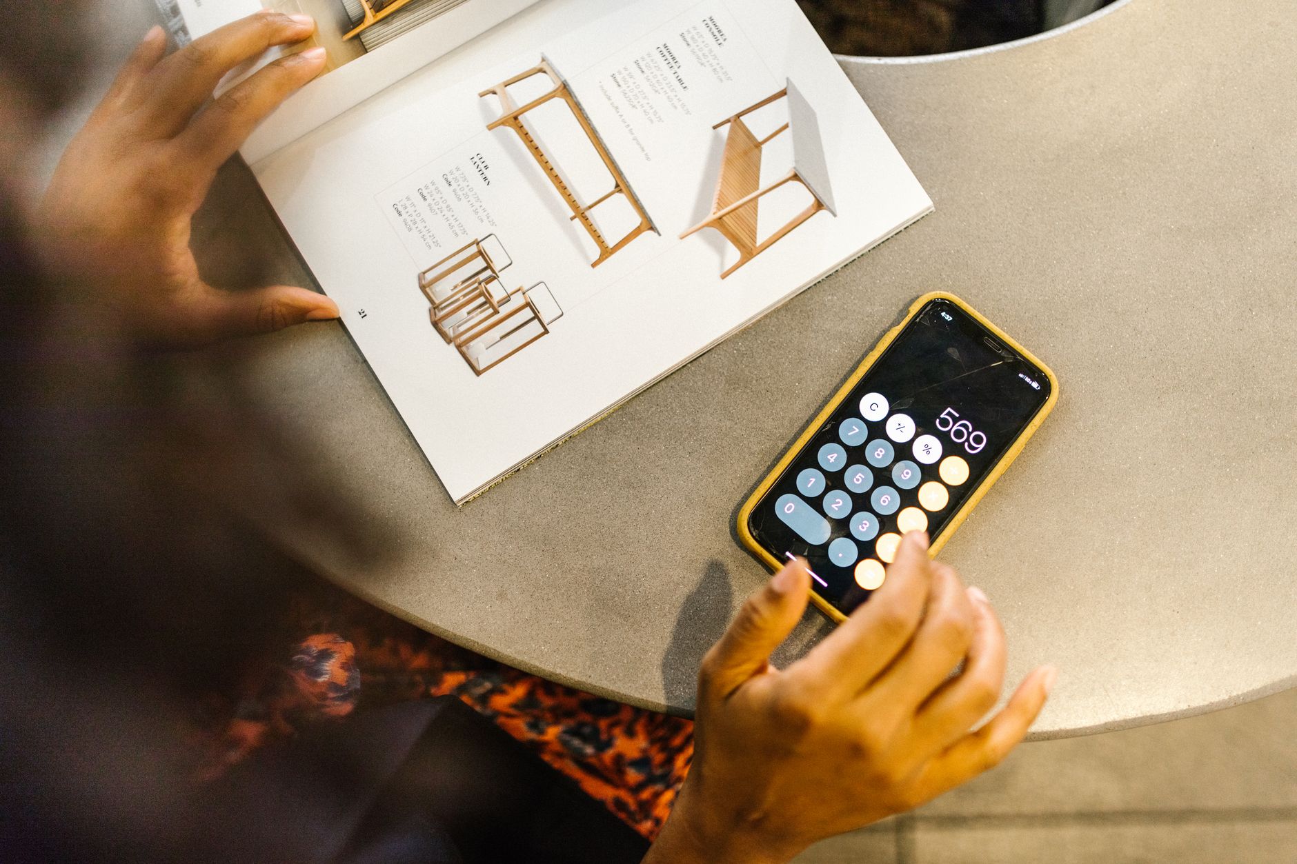 Une personne utilise la calculatrice d’un smartphone à coque jaune près d’un catalogue de meubles ouvert, sur une table grise.