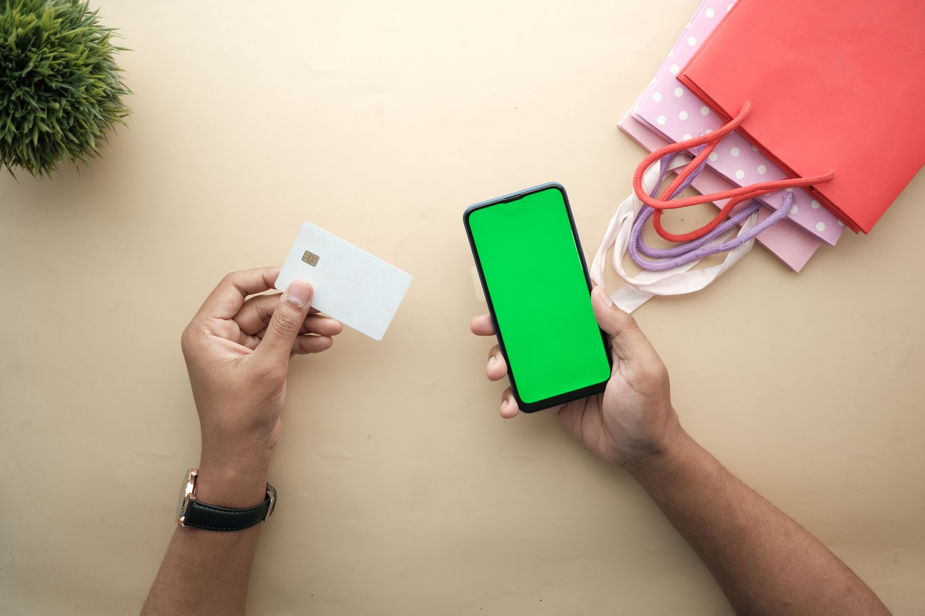 Vue de dessus de deux mains tenant une carte bancaire blanche et un smartphone à écran vert, avec des sacs de shopping et une plante sur une table beige.